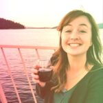 A woman enjoying a glass of red wine on a boat during sunset with a scenic water and forest background.