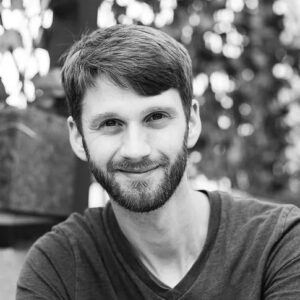A smiling young man with a beard and short hair, outdoors with blurred trees in the background, representing professionalism and approachability.